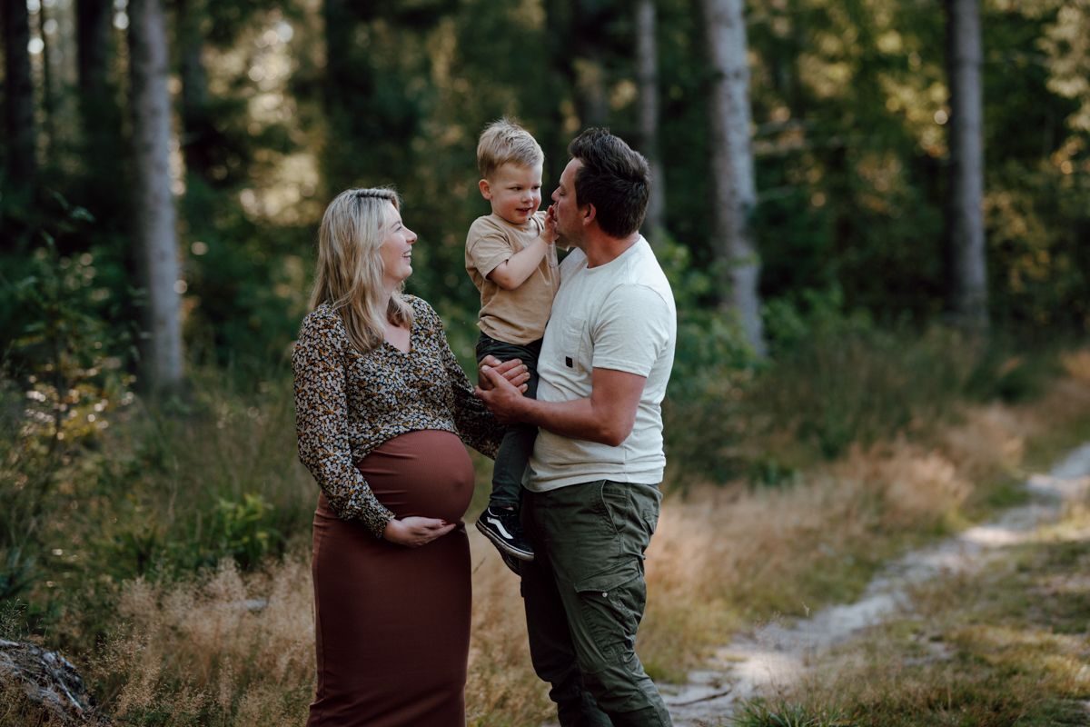 Een zwanger gezin staat op een bospad; de ouders houden een peuter vast en naast hen staat een zwangere vrouw die glimlacht.