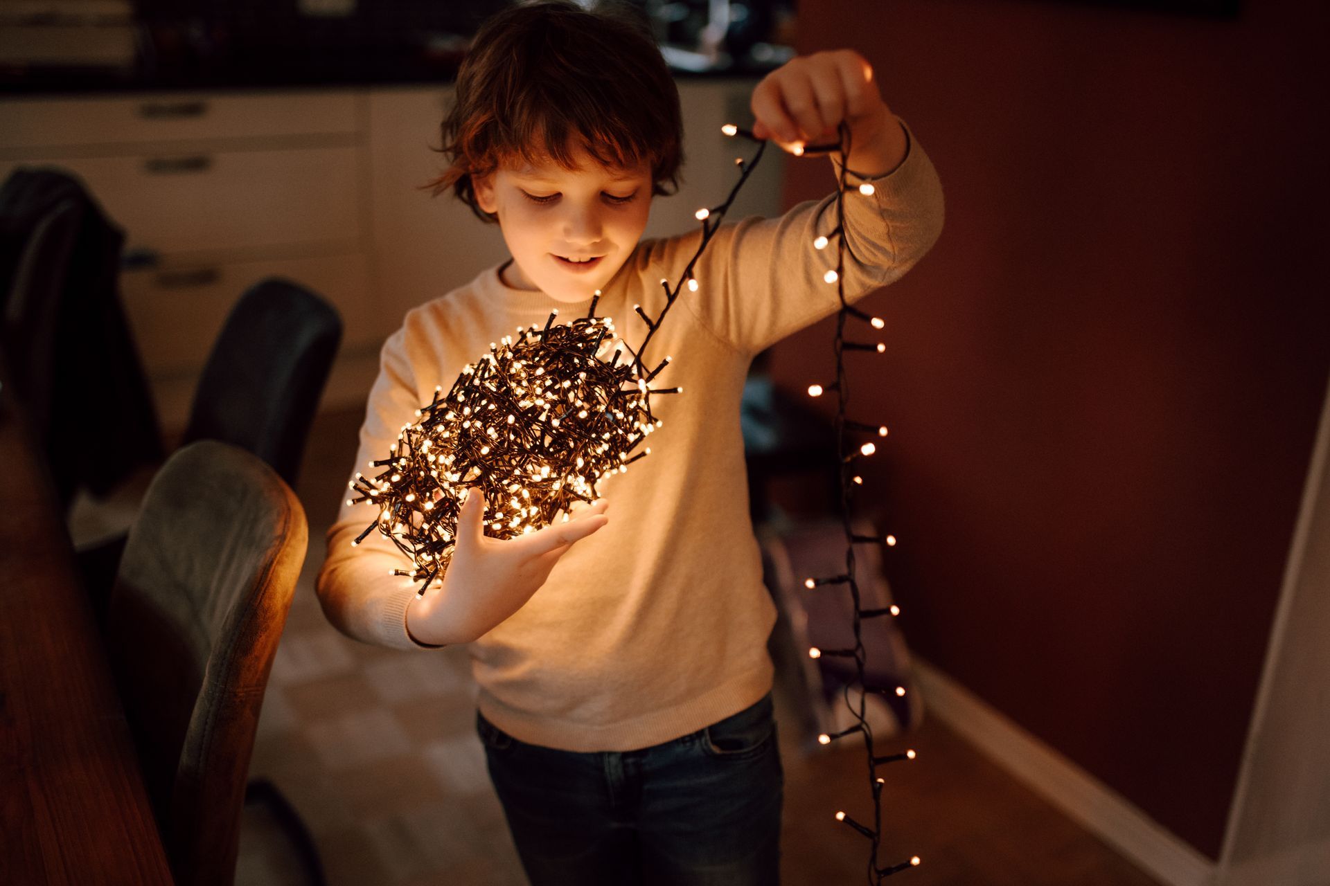 Een persoon in een lichtgekleurde trui houdt een gloeiende, in elkaar verstrengelde bol van warmwitte lichtsnoeren vast in een schemerig verlichte ruimte.