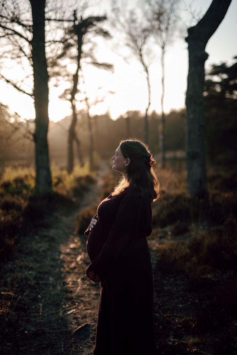 Silhouetted pregnant person standing on a forest path at sunset, backlit by warm light