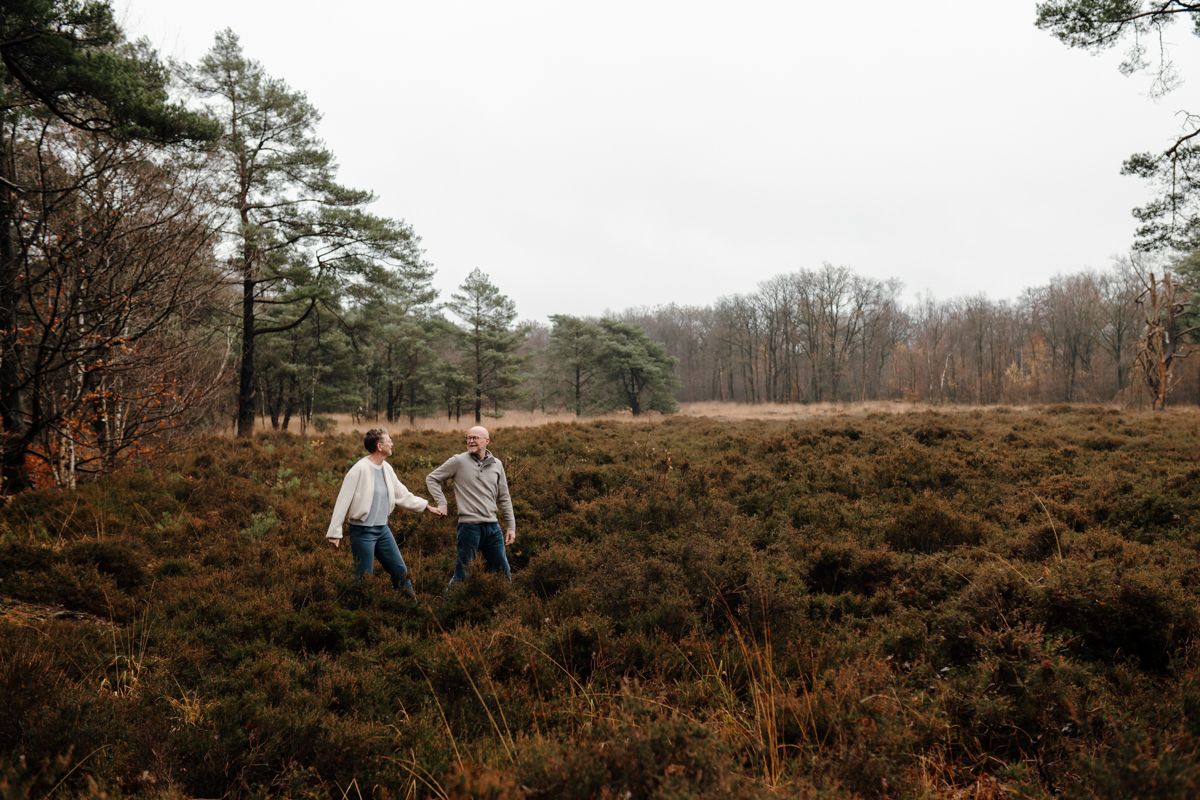 Twee mensen lopen hand in hand door een veld met laaghangende struiken, met op de achtergrond een bosrand. Tijdens een fotoshoot