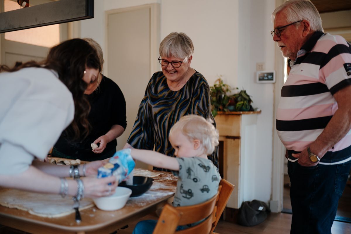 Een groep mensen verzamelt zich rond een keukentafel en helpt een peuter met het strooien van bloem over deeg, terwijl ze samen eten klaarmaken.