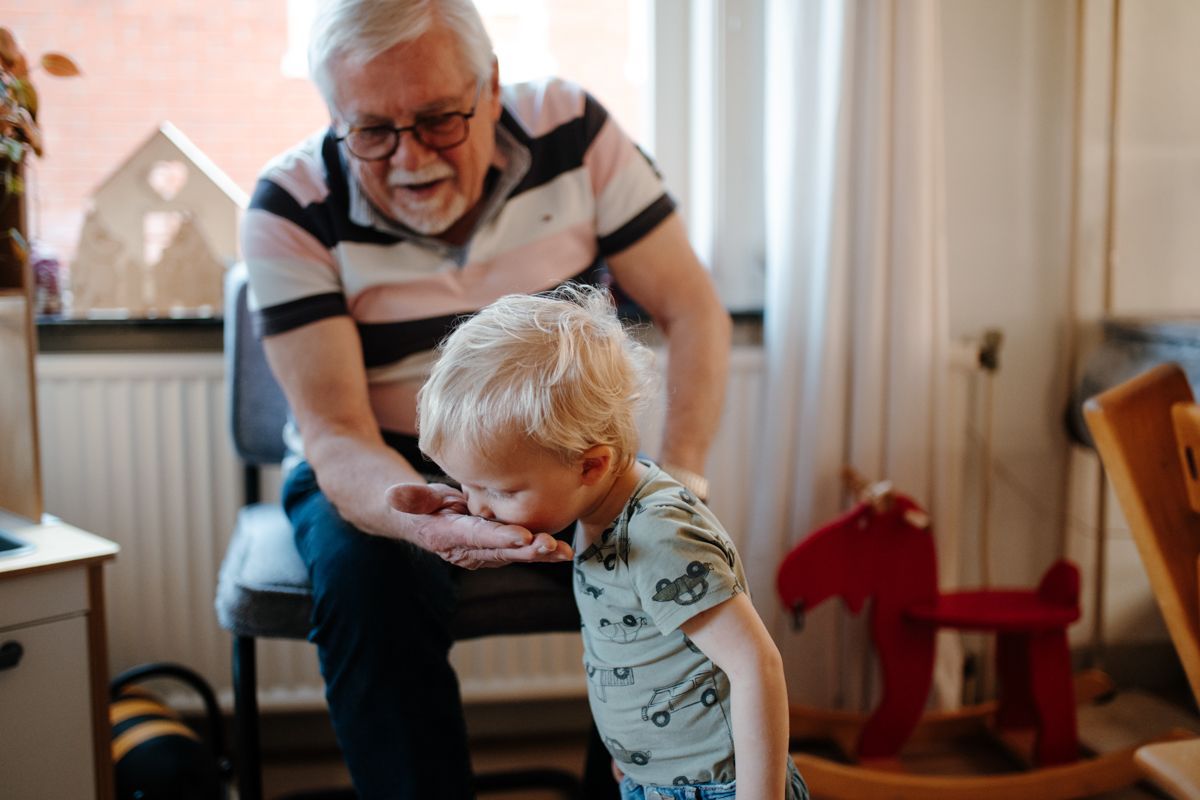 Een kind buigt zich voorover om uit de hand van een volwassene te eten, terwijl ze samen in een kamer zitten met een speelgoedhobbelpaard in de buurt.