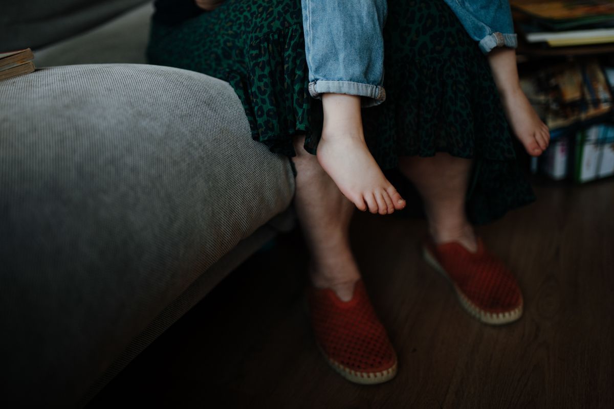 Een persoon, gekleed in een groene rok met patroon en rode instapschoenen, zit met een kind op schoot, waarbij de voetjes bijna de grond raken.