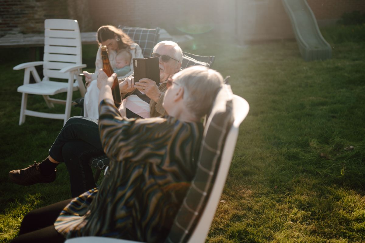 Drie mensen ontspannen in een grasrijke achtertuin tijdens zonsondergang, waarbij een van hen een glazen fles tegen het licht houdt.
