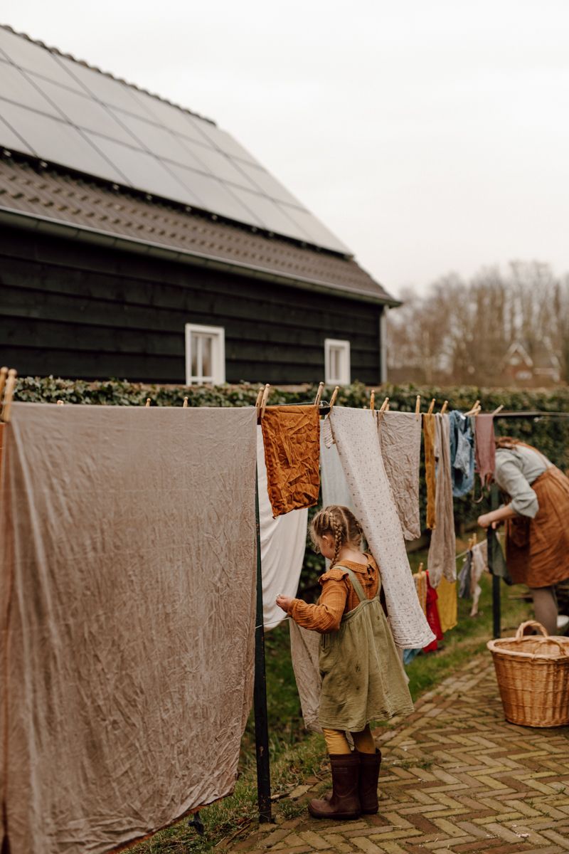 Een kind en een volwassene hangen de was op aan een waslijn buiten, voor een donkere schuur met zonnepanelen op het dak.