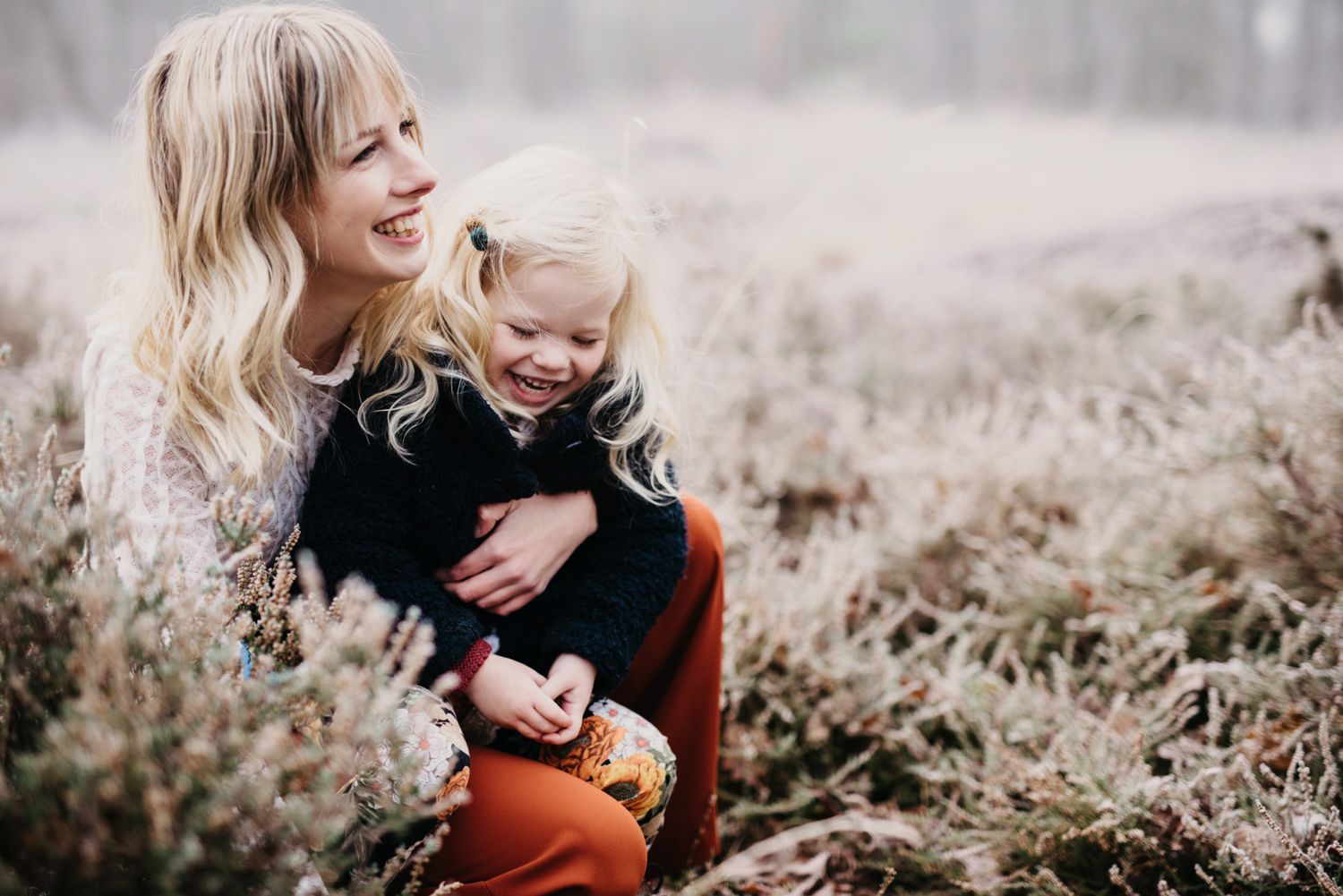 Twee lachende kinderen zitten in het hoge gras, de ene knuffelt de andere buiten.