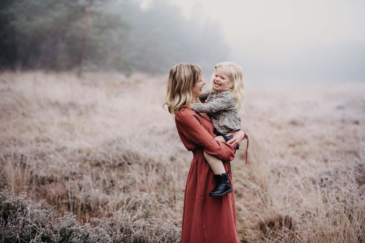 Een vrouw in een rode jurk houdt een kind vast in een bevroren veld, tegenover elkaar in een mistig landschap.