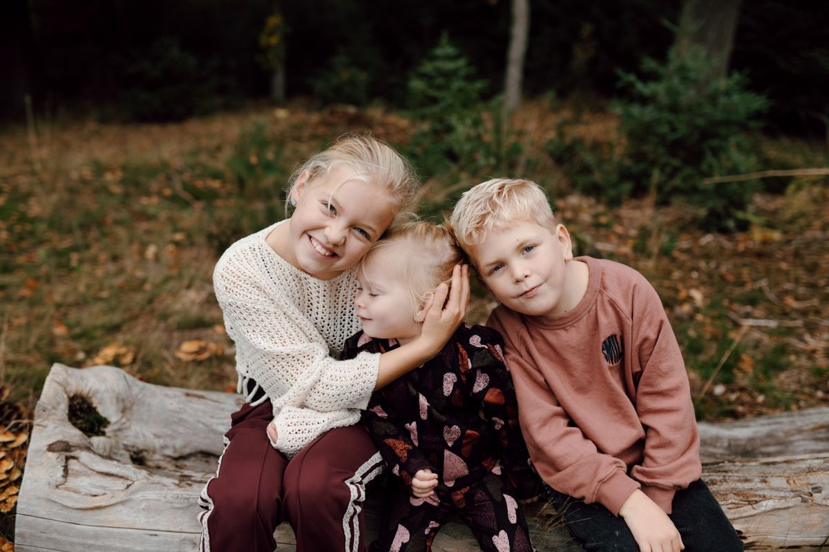 Drie kinderen zitten buiten op een boomstam, tegen elkaar aan geleund en lachend.