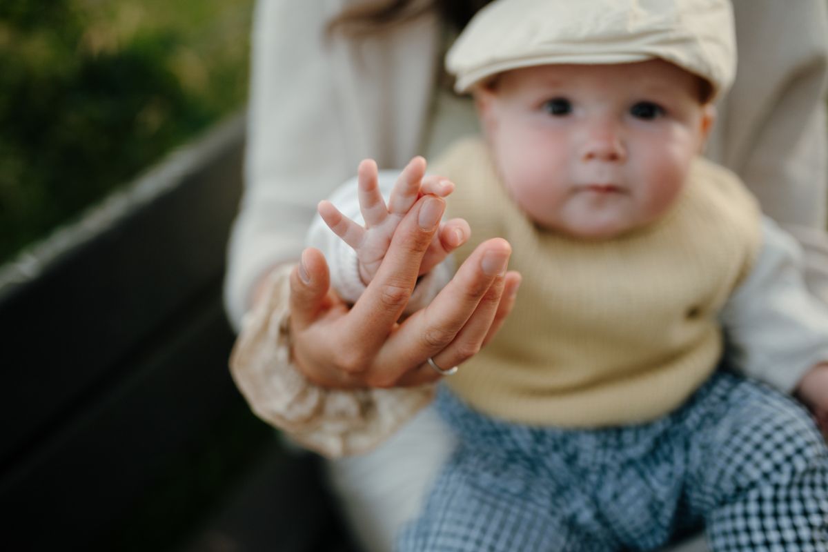 Een volwassene houdt buiten de hand van een baby vast; de baby draagt een beige mutsje en een trui.