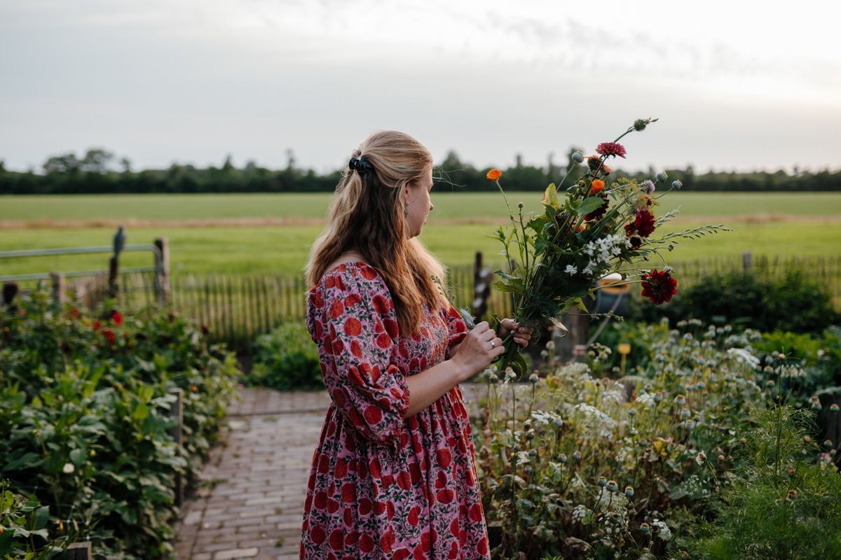 Een persoon in een rode jurk met bloemenprint houdt een boeket wilde bloemen vast terwijl ze bij schemering in een tuin met bakstenen paden staat.