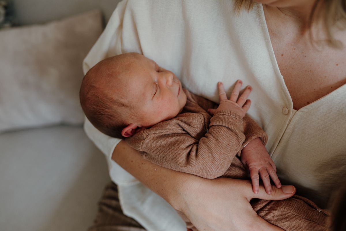 Een slapende baby in een bruin gebreid pakje ligt in de armen van een volwassene op een bank.