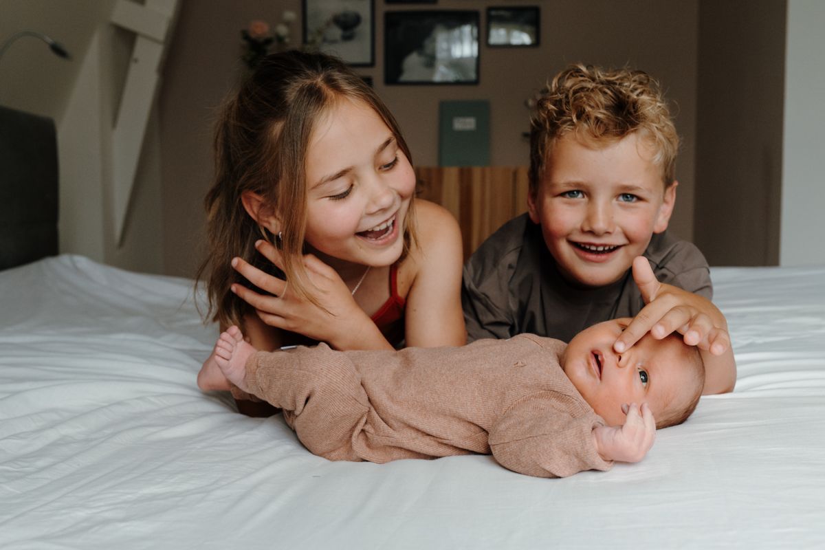Twee lachende kinderen liggen op een bed met een pasgeboren baby, gewikkeld in een beige deken.