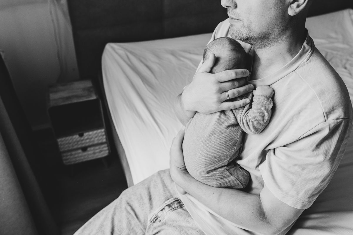 Een man houdt een slapende baby vast op een bed, op een zwart-witfoto.