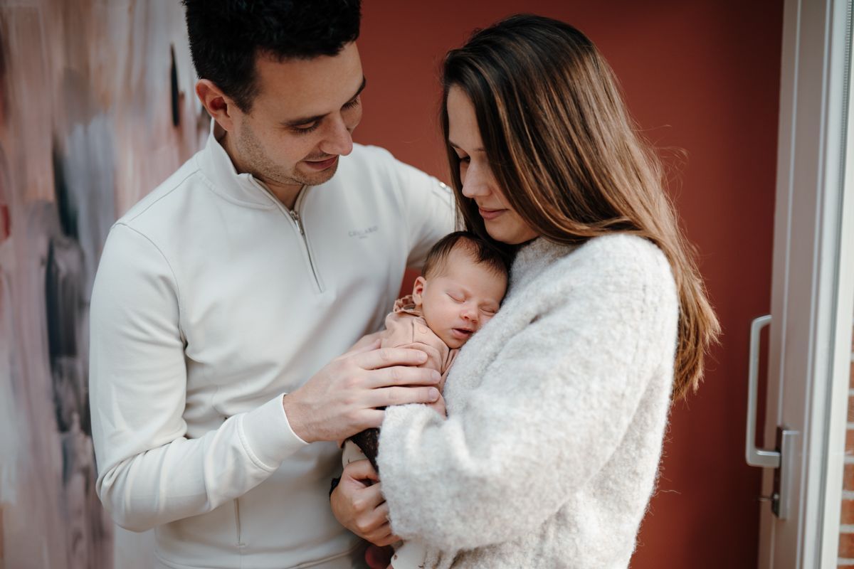 Twee volwassenen houden een pasgeboren baby voorzichtig vast in een deuropening, glimlachend in een warme binnenruimte.