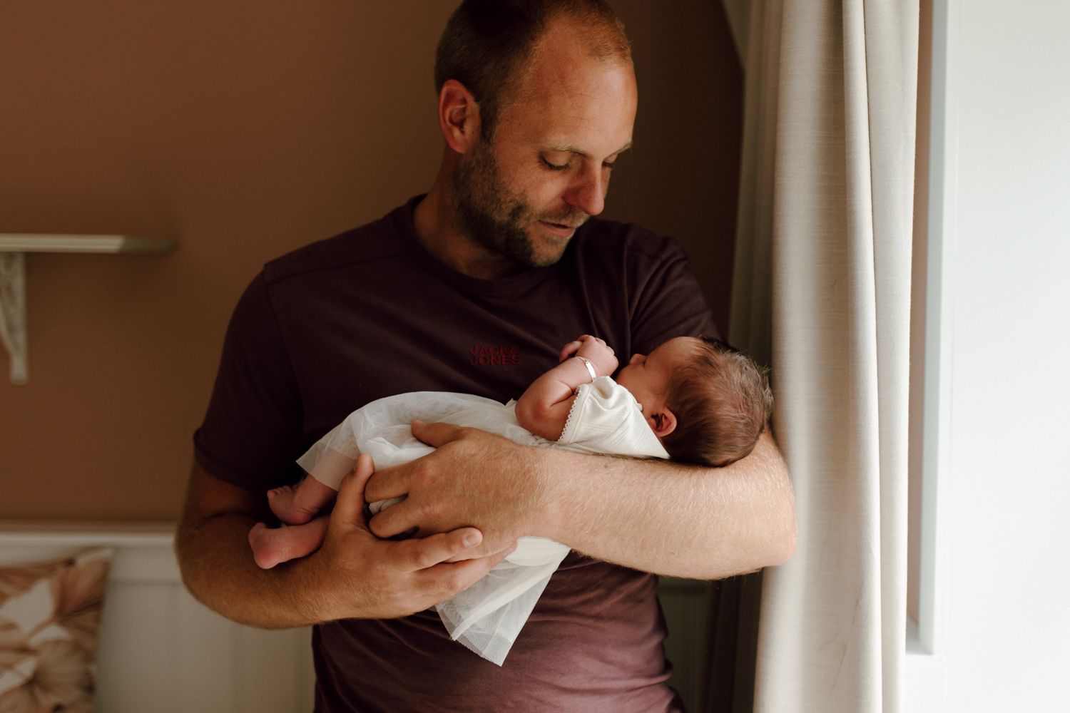 Een man houdt een ingewikkelde baby vast bij een raam en kijkt liefdevol naar beneden.