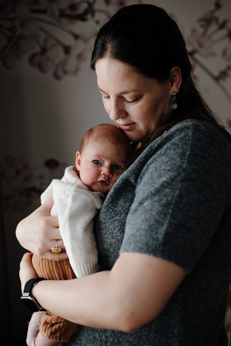 Een vrouw houdt een baby vast die in een witte deken is gewikkeld; beiden kijken naar beneden in een zacht verlichte binnenruimte.