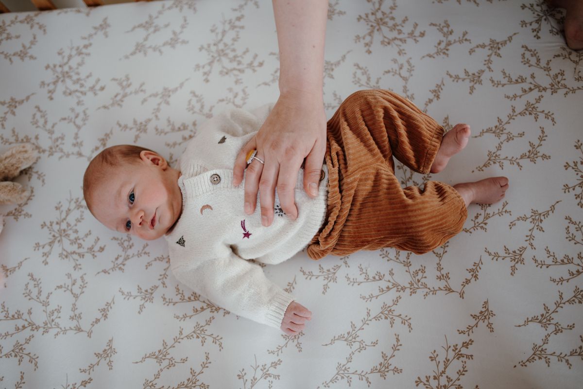 Een baby ligt op een dekentje met bloemenprint en kijkt omhoog, terwijl een volwassen hand op de borst rust.