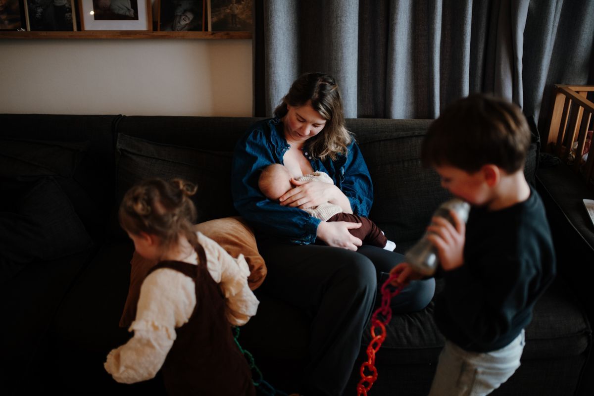 Moeder voed haar baby terwijl haar andere twee kinderen door de kamer lopen met een fles en speelgoe