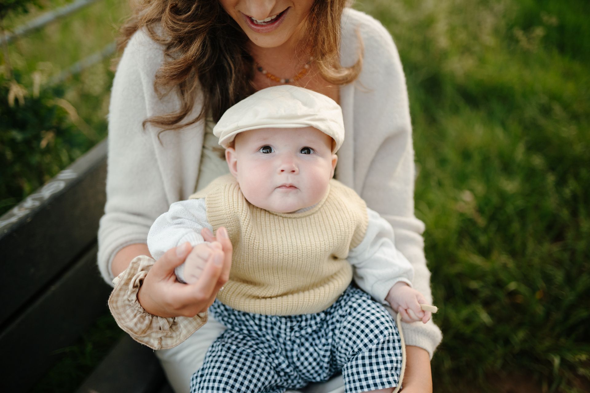 A baby wearing a cream vest and newsboy cap sits on a person's lap on a wooden bench outdoors.