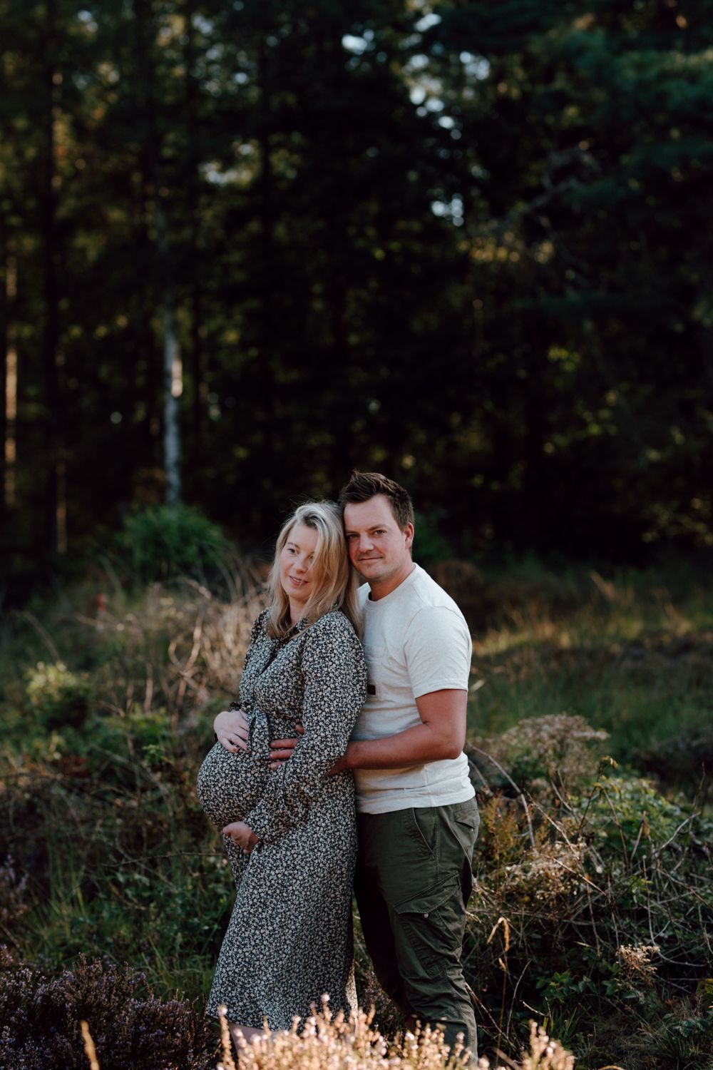 Zwangerschapsfotografie buiten Een stel dat elkaar omhelst op een open plek in het bos bij schemering, omringd door bomen en gras.