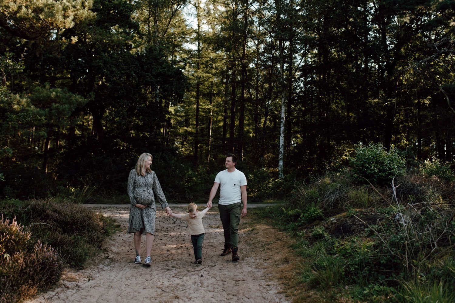 Zwangerschapsfotografie buiten Drie mensen die hand in hand lopen over een bospad, omringd door bomen en groen.