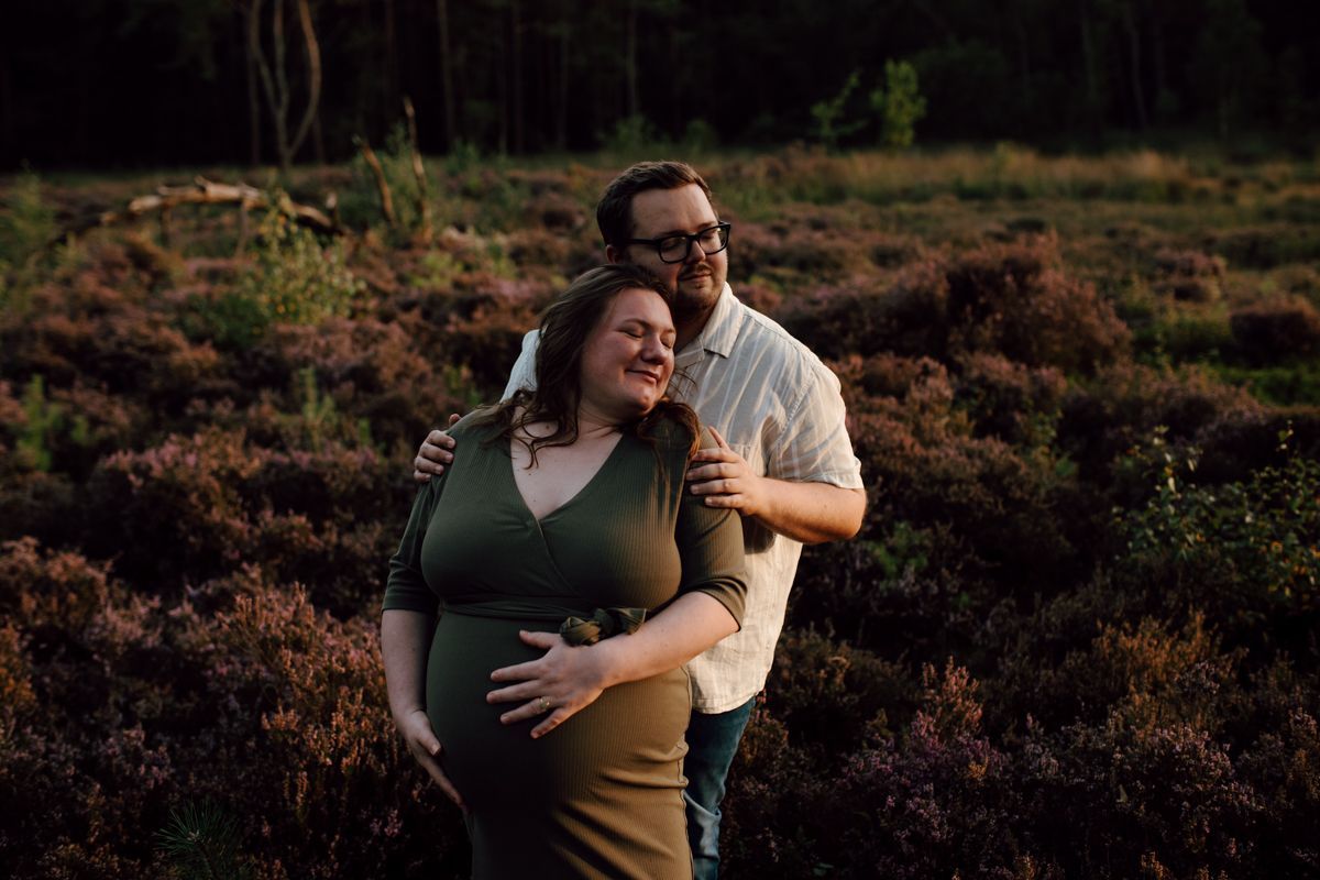 Een stelletje dat elkaar omhelst in een veld bij zonsondergang, glimlachend met het bos op de achtergrond.