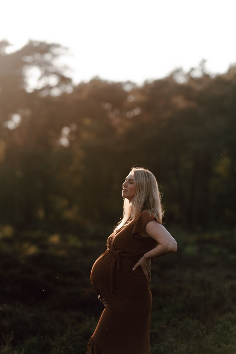 Een zwangere vrouw in een bruine jurk staat buiten bij zonsondergang, met bomen op de achtergrond.