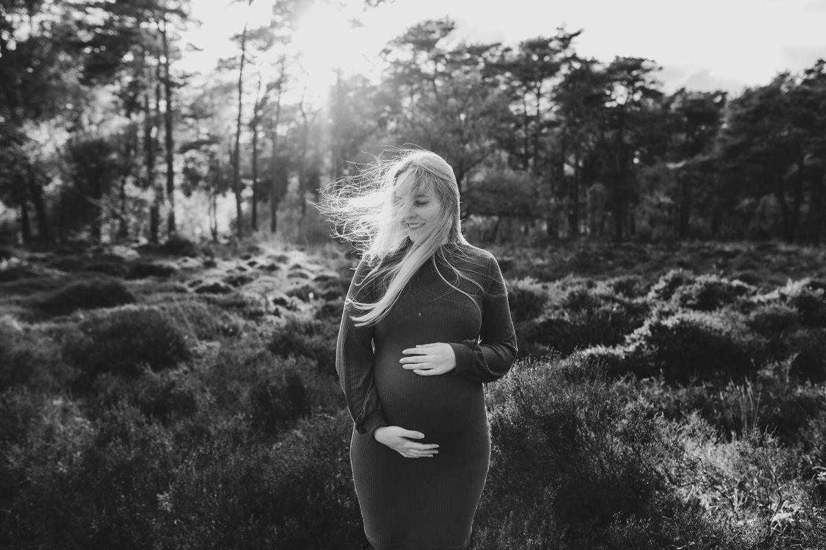 Een zwangere vrouw staat in een zonovergoten bos tijdens haar zwangerschapsshoot, met haar handen op haar buik, haar haar wapperend in de wind en donkere kleding.