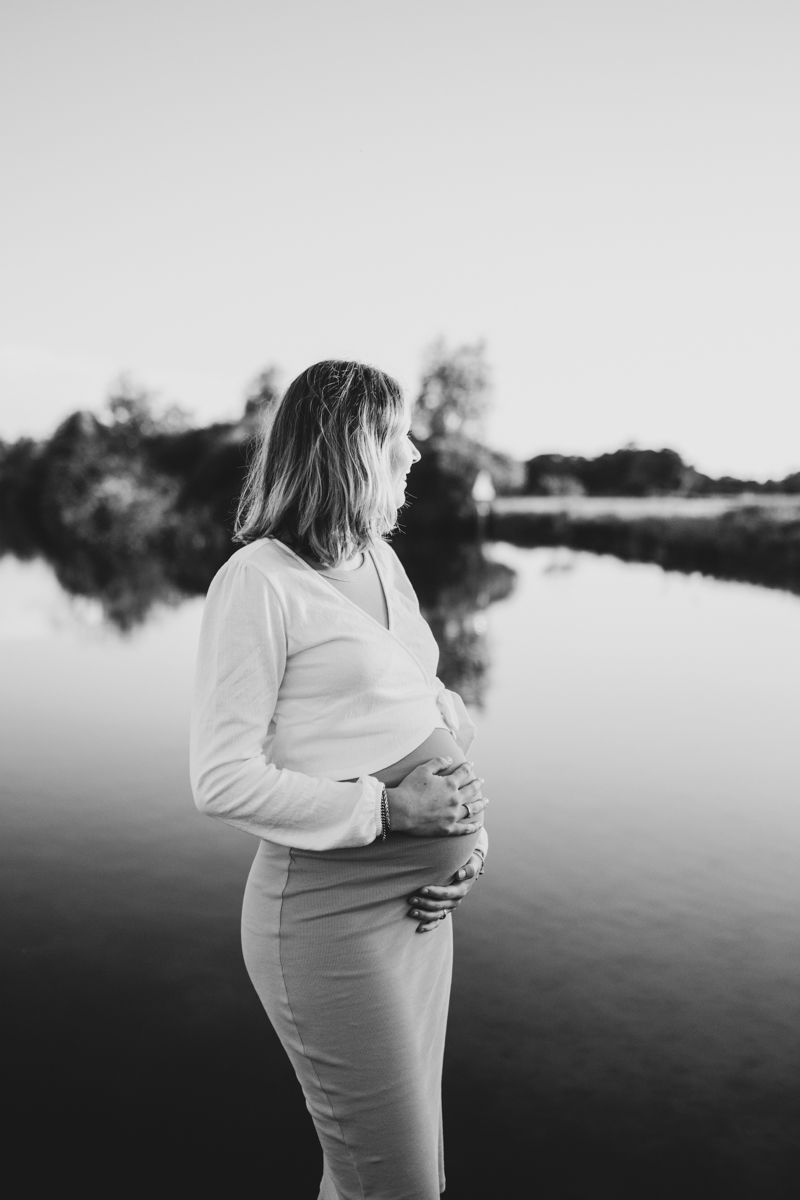Een zwangere vrouw in een witte top en broek staat bij een rustig meer (zwart-wit).