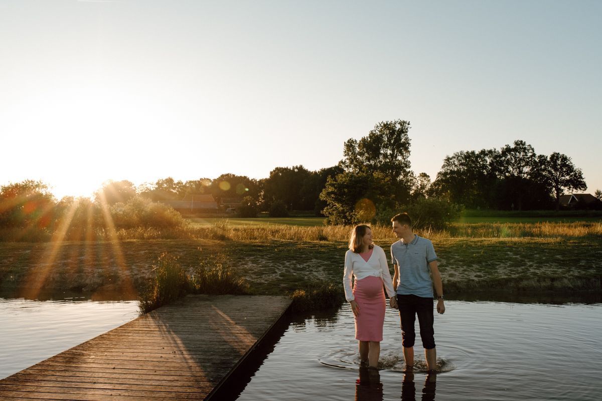 Twee mensen staan op een steiger bij zonsondergang, aan de rand van kalm water en tussen de wilde bloemen.