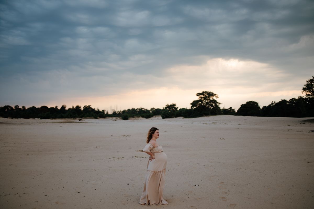 Zwangere vrouw op open zandvlakte onder dramatische avondlucht, zwangerschapsfotograaf Friesland en Drenthe