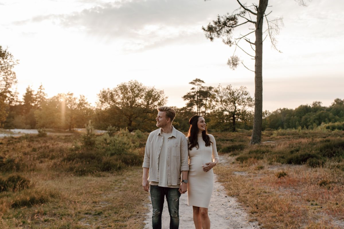 Een zwanger stel loopt bij zonsondergang over een grindpad in een grasveld tijdens een fotoshoot