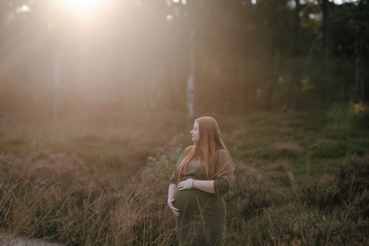 Een zwangere vrouw staat in een zonovergoten grasveld tijdens haar zwangerschap fotoshoot en kijkt opzij met de armen over elkaar.