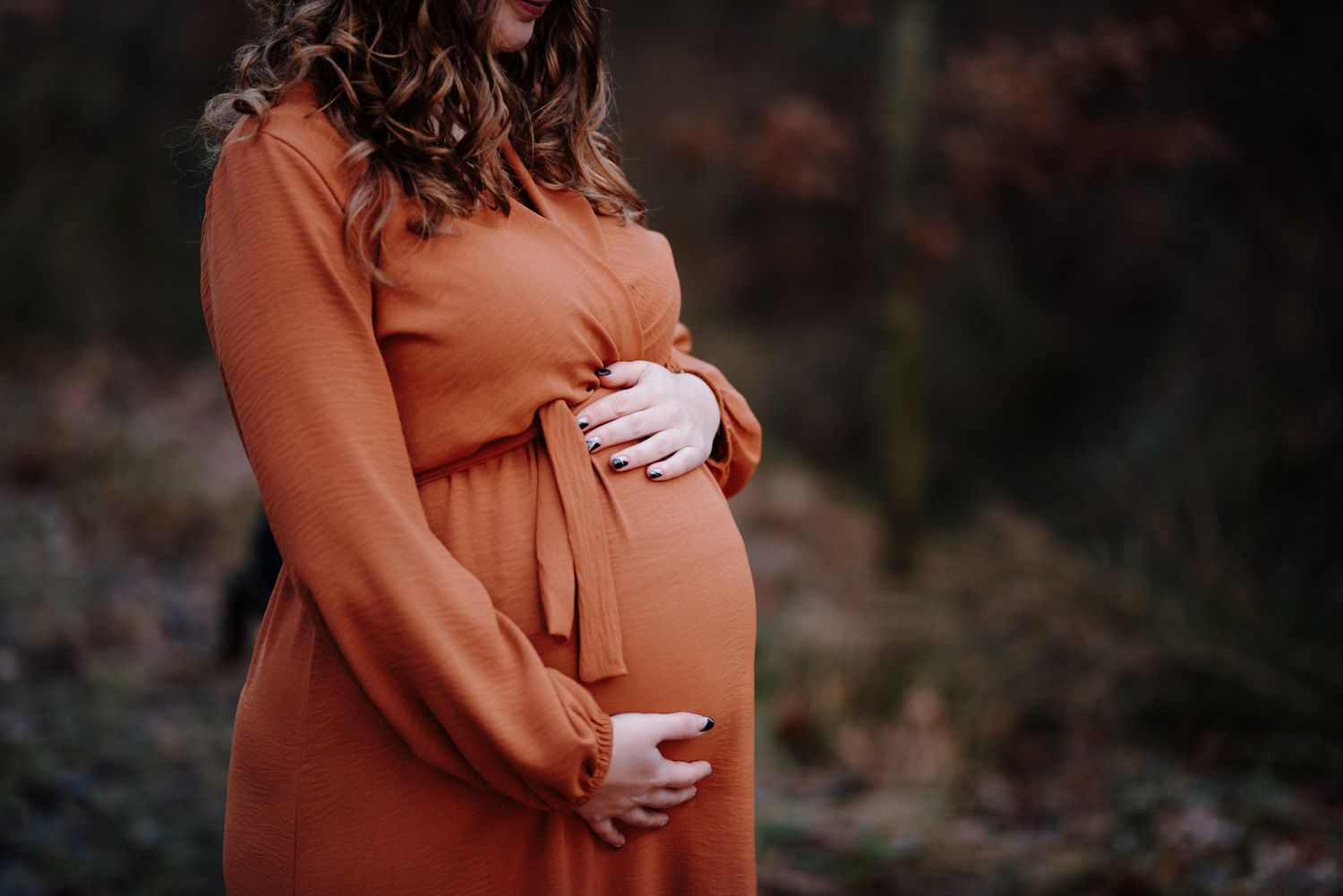 Een vrouw in een oranje jurk houdt haar zwangere buik vast terwijl ze in een bosrijk gebied staat.