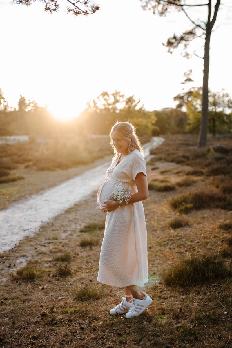 Een zwangere vrouw in een witte jurk staat op een zonovergoten pad bij zonsondergang.