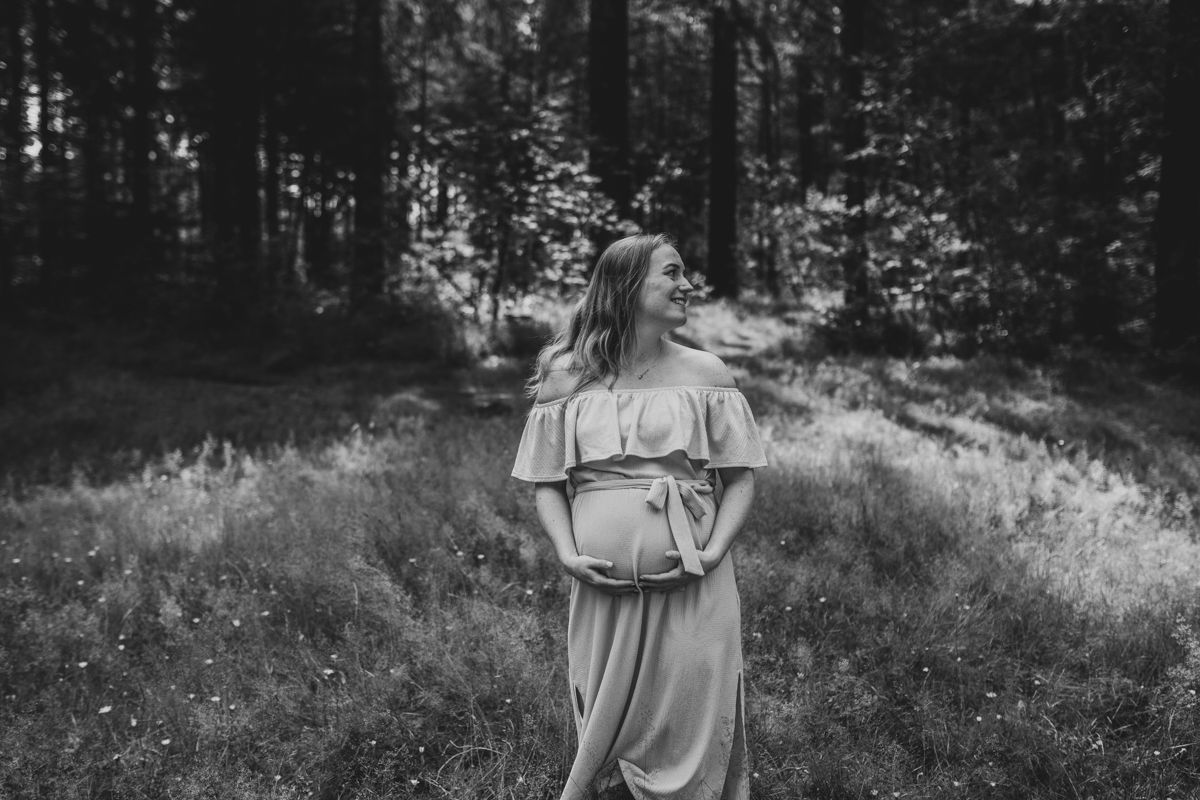 Vrouw in een zwierige jurk staat op een open plek in het bos en kijkt opzij tijdens de fotoshoot van haar zwangerschap