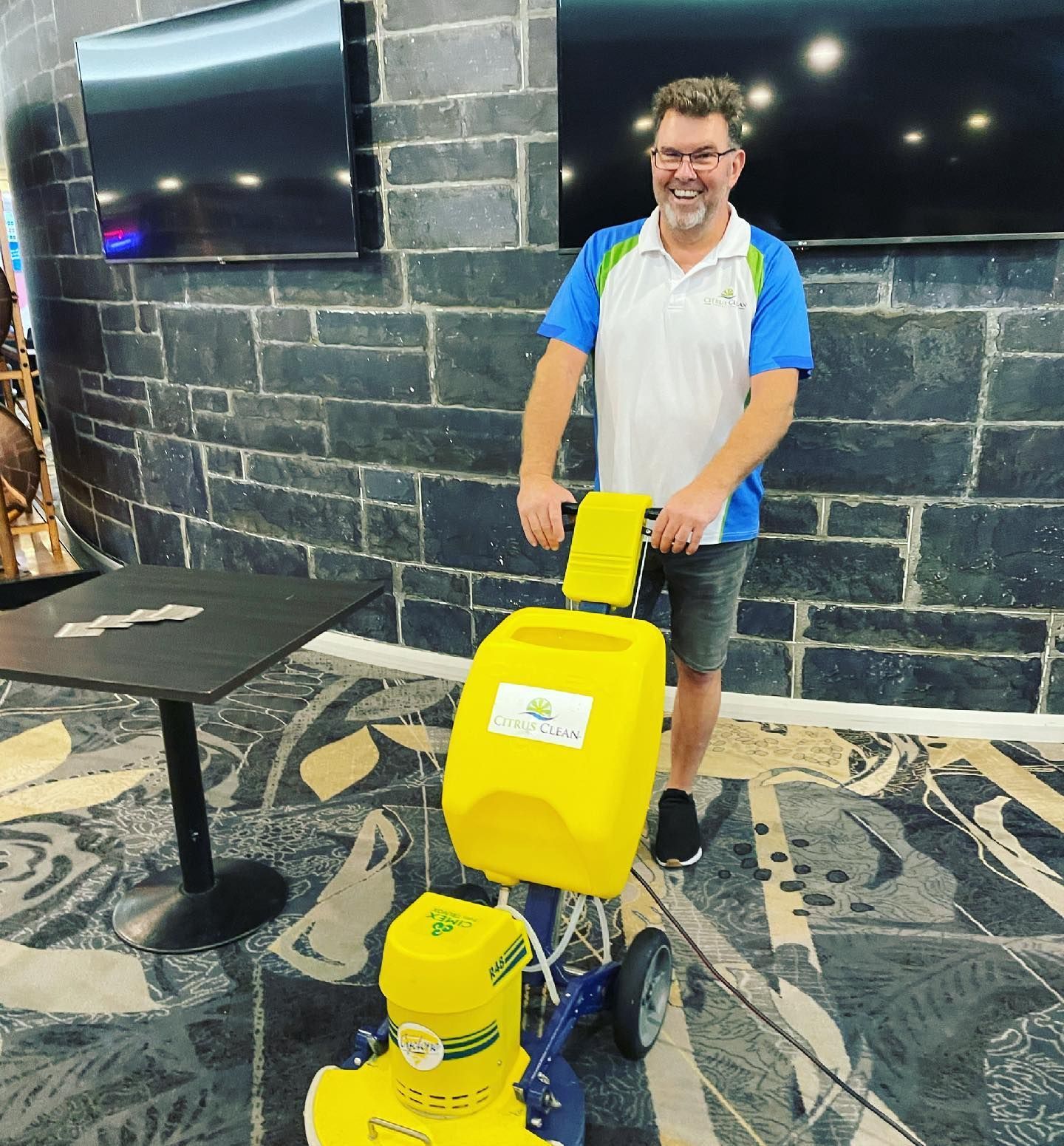 A smiling person in a blue and white polo shirt operates a bright yellow floor cleaning machine indoors.