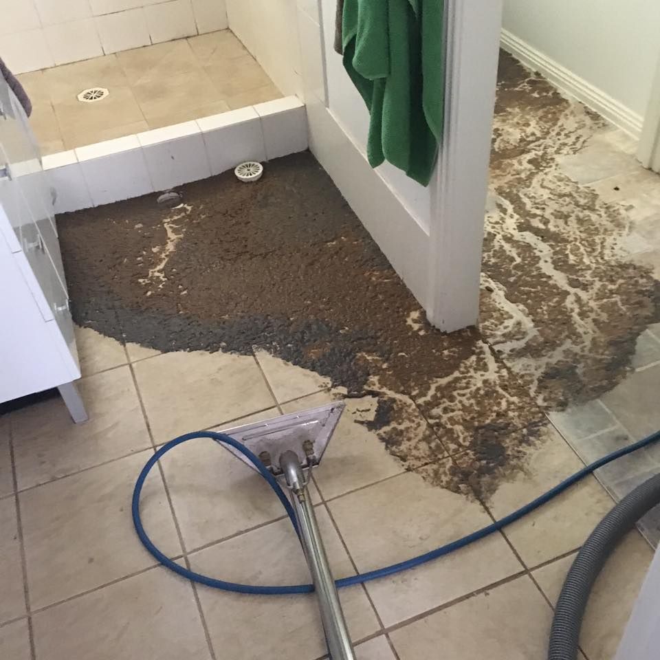 A professional tile cleaning wand sits on a tiled floor being cleaned of a large, dark spill spreading toward a shower.
