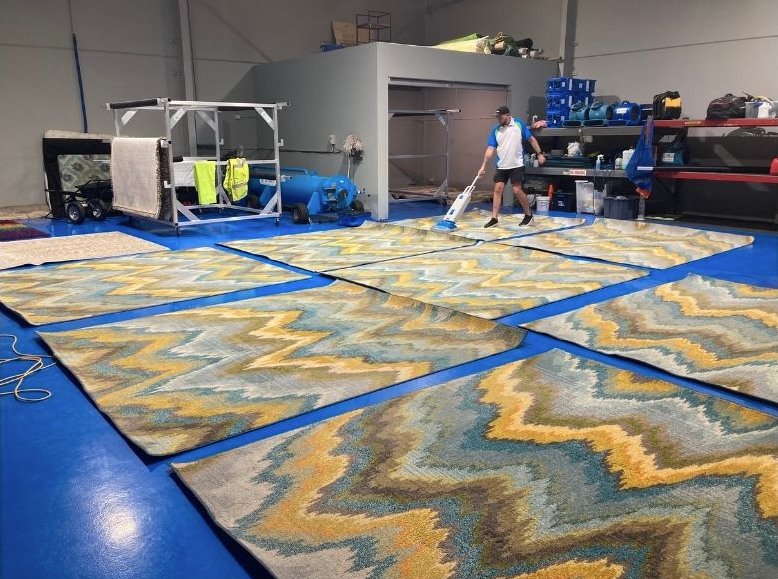 A worker vacuums several large, chevron-patterned blue and yellow rugs laid out on the blue floor of a warehouse.