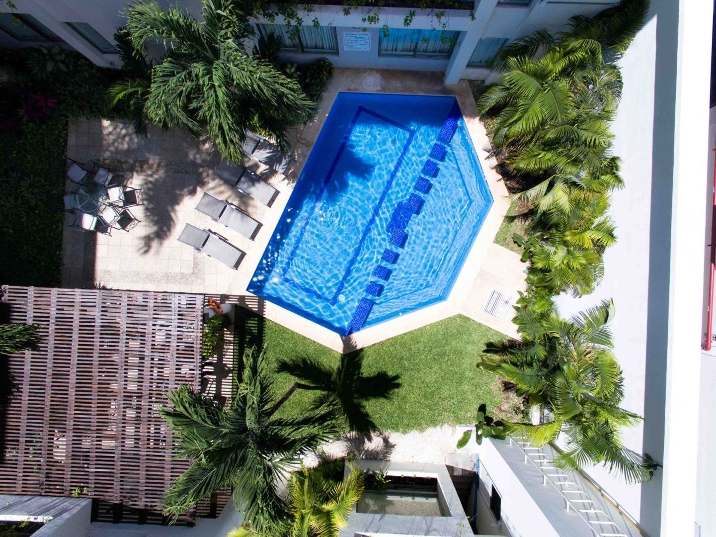 Vista aérea de una piscina geométrica azul rodeada de jardines, muebles de patio y edificios.