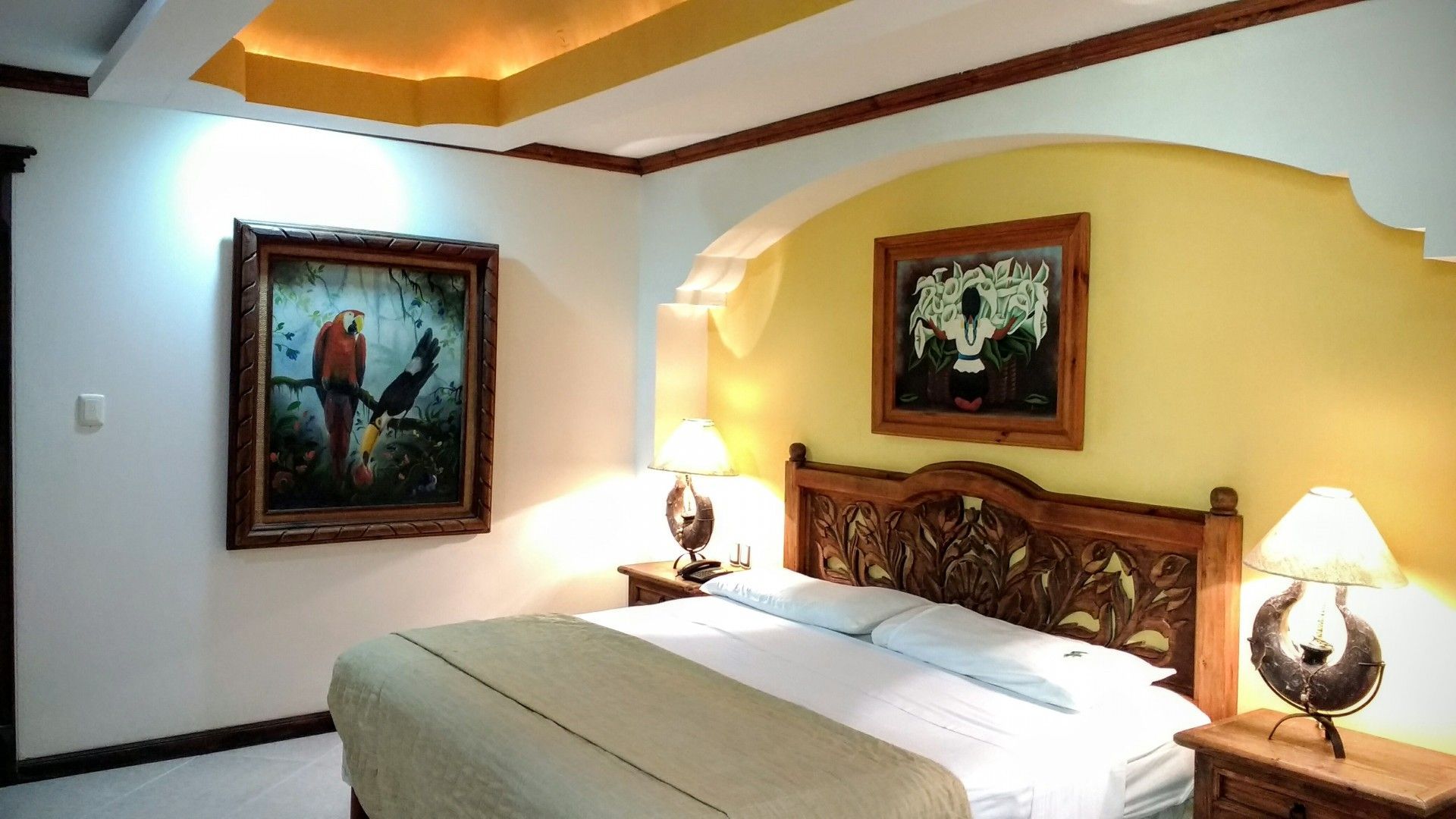 Dormitorio con cama, mesitas de noche, lámparas, obras de arte y puerta arqueada; colores blanco, amarillo y marrón.