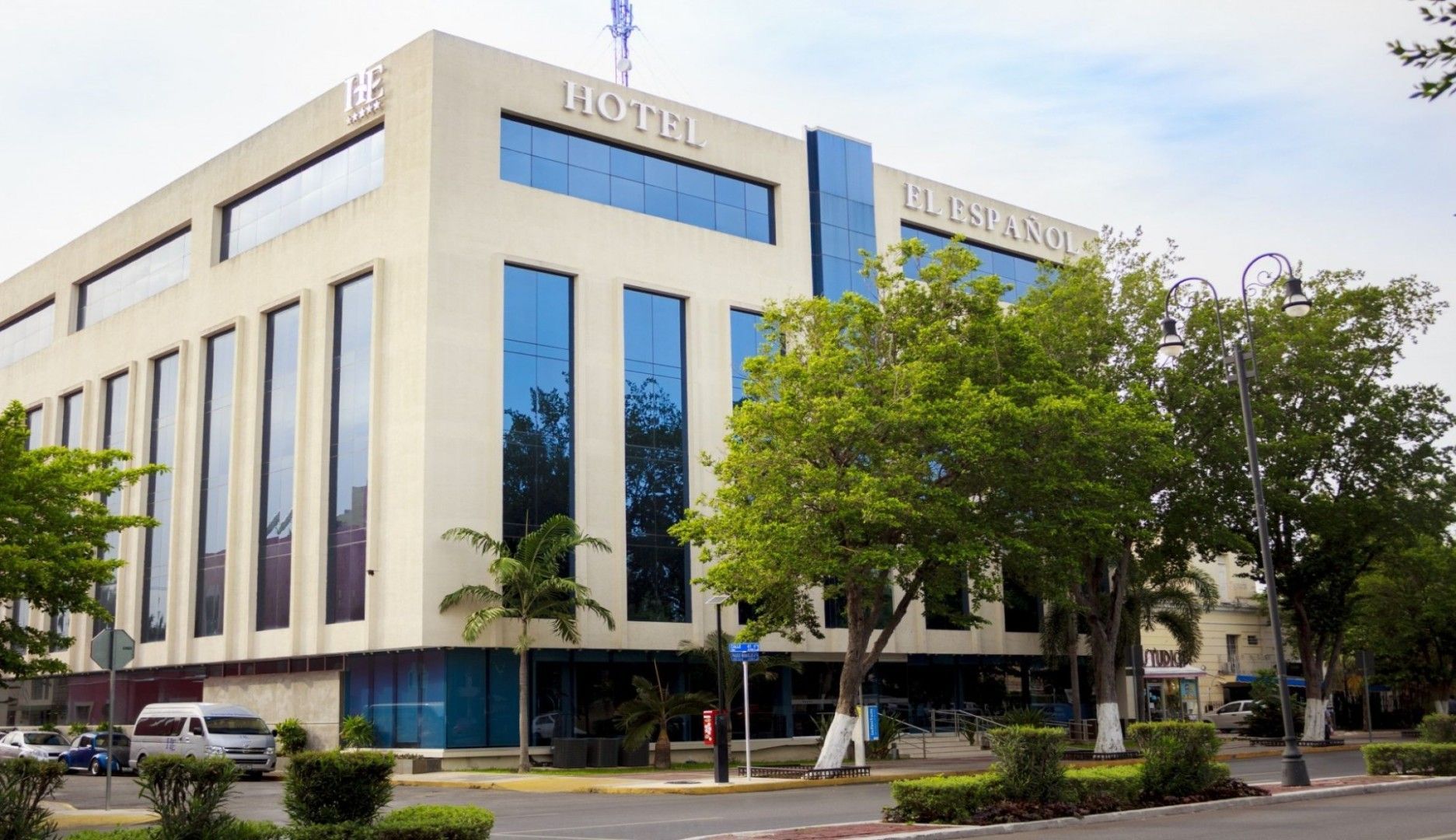 Exterior del hotel con altos ventanales azules, fachada blanca y árboles circundantes.