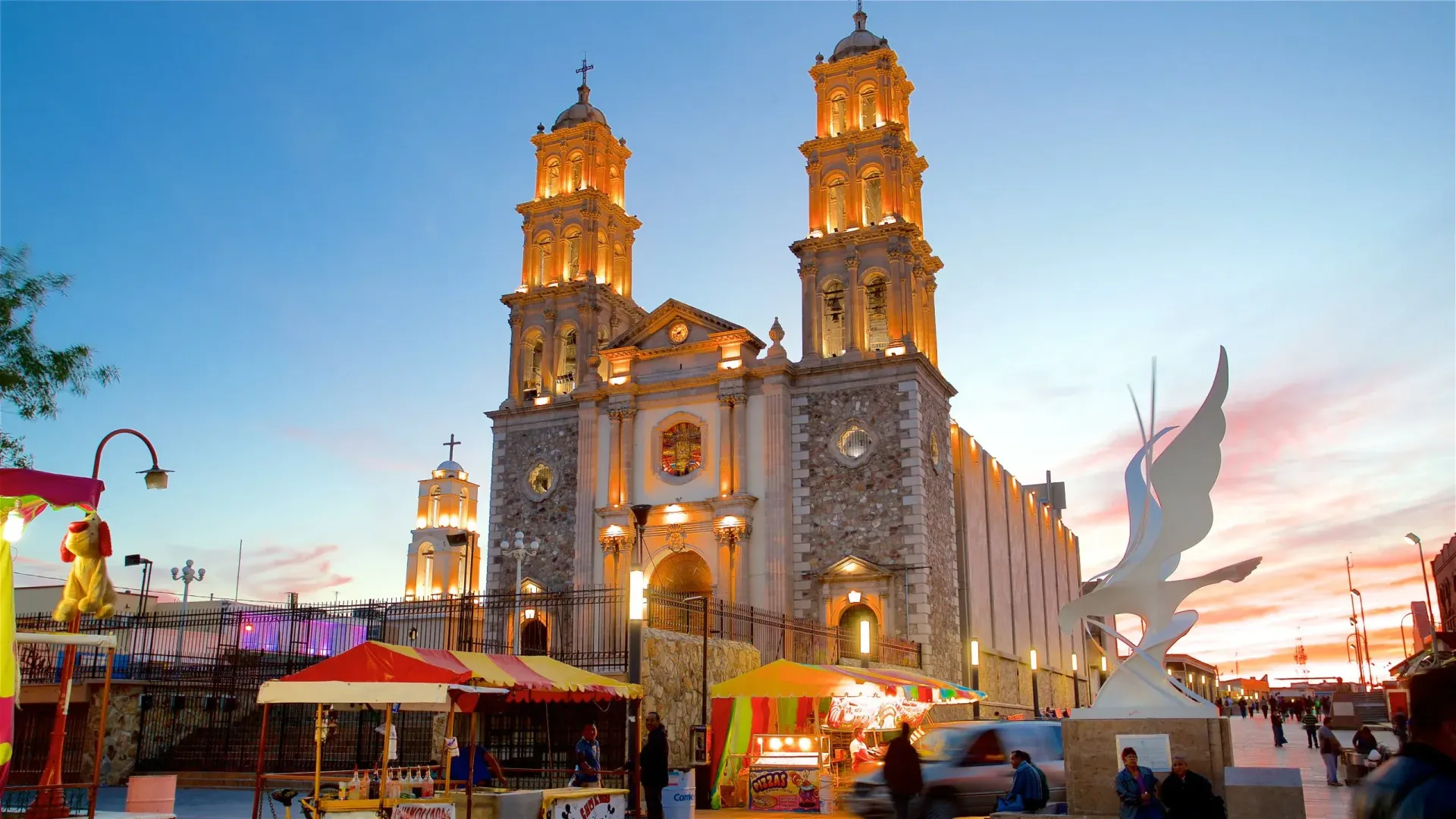 Iglesia iluminada al anochecer, con puestos de comida y esculturas en primer plano.