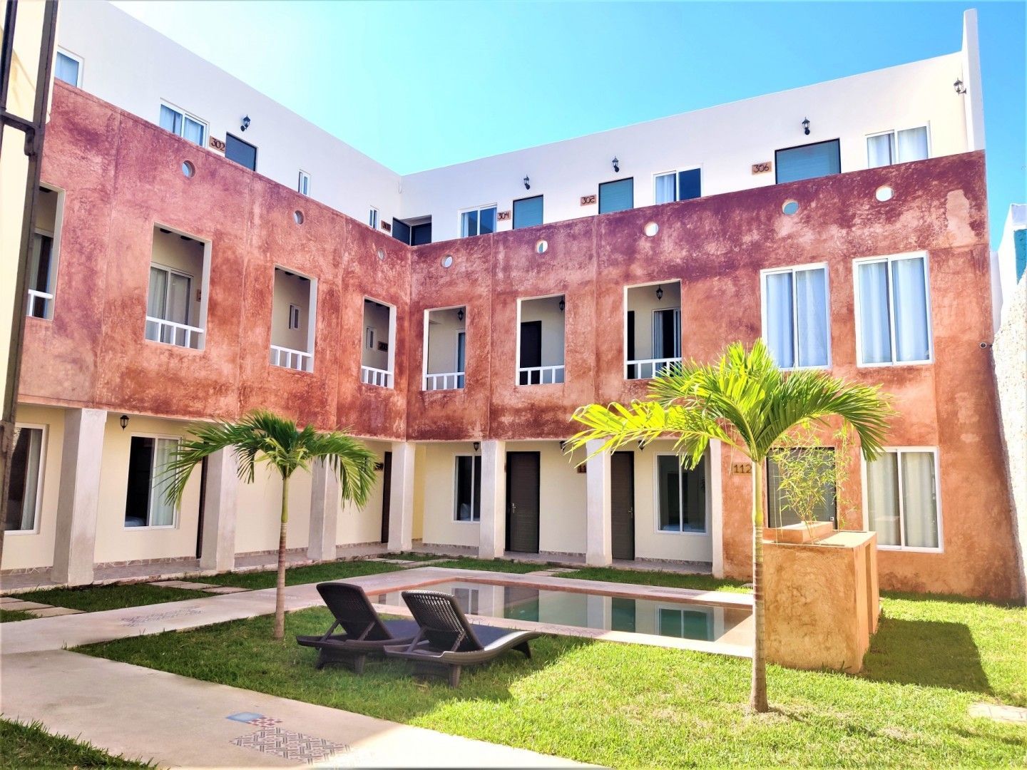Hotel con patio, piscina, césped, palmeras y edificio de varios pisos con ventanas.