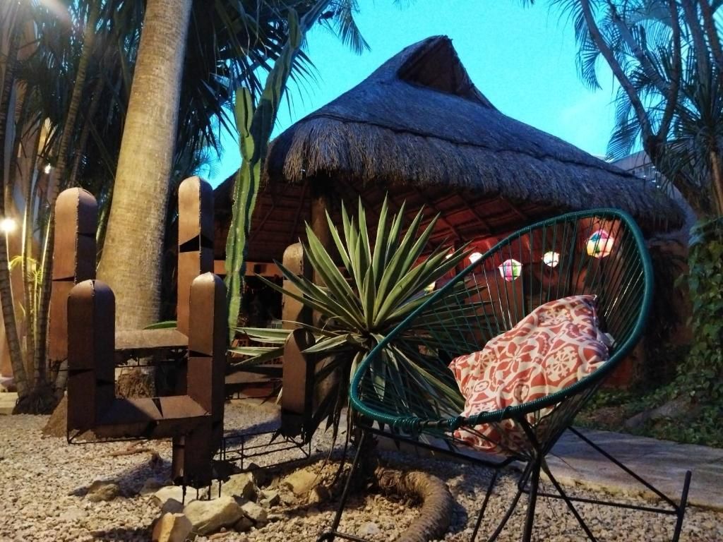 Una estructura con techo de paja con una silla verde, una escultura de cactus de metal y plantas.