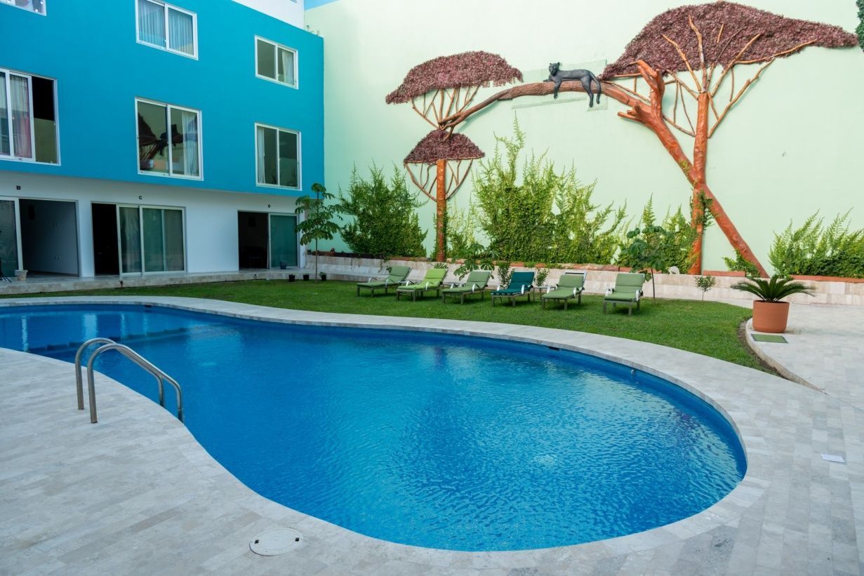 Área de piscina con agua azul, césped verde, camastros y un edificio turquesa.