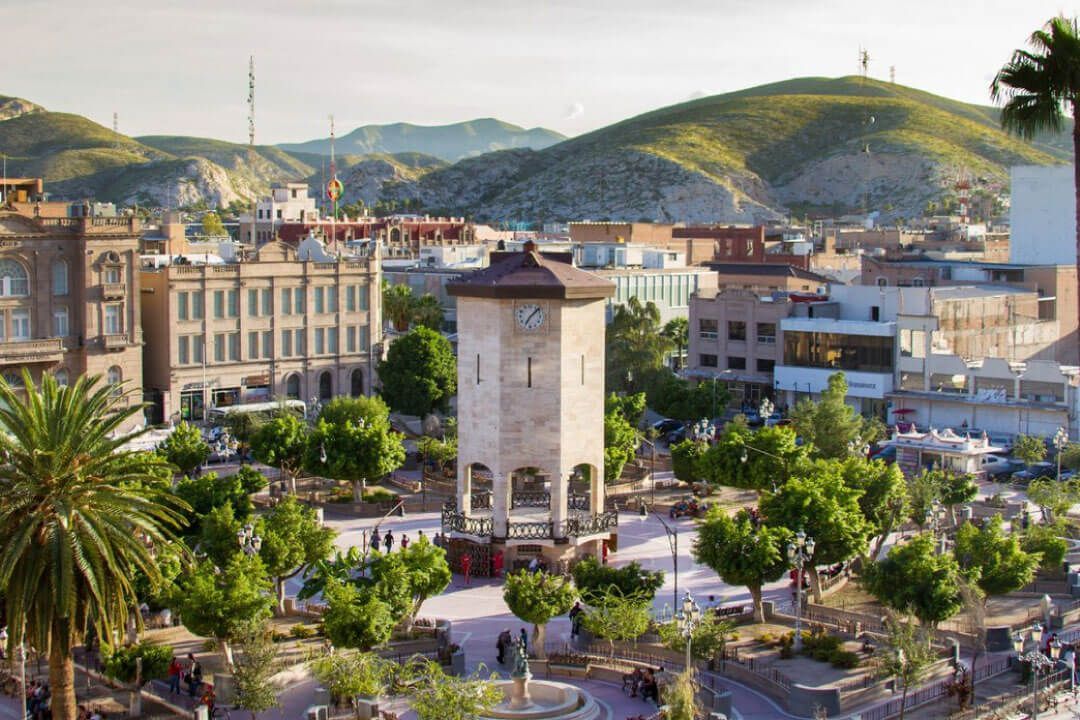 Plaza de la ciudad con torre del reloj, árboles, edificios y colinas.