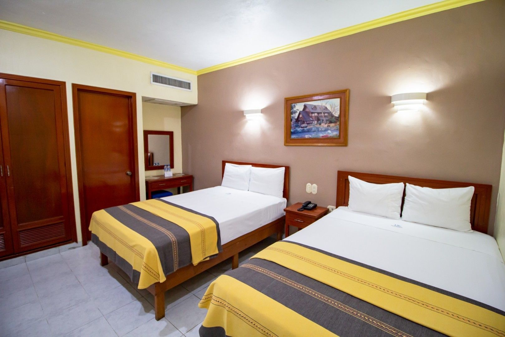 Habitación de hotel con dos camas, mantas marrones y amarillas, obras de arte y una pequeña mesa.