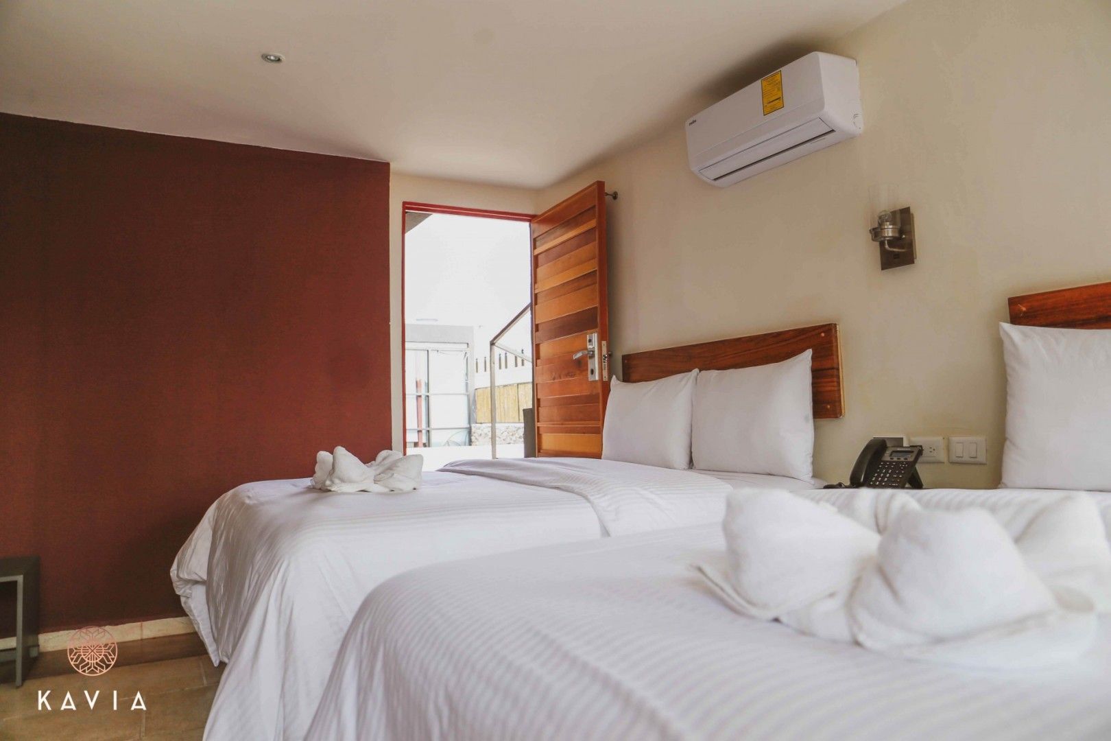 Habitación de hotel con dos camas, puerta abierta, pared con acento rojo, ropa de cama blanca y unidad de aire acondicionado.