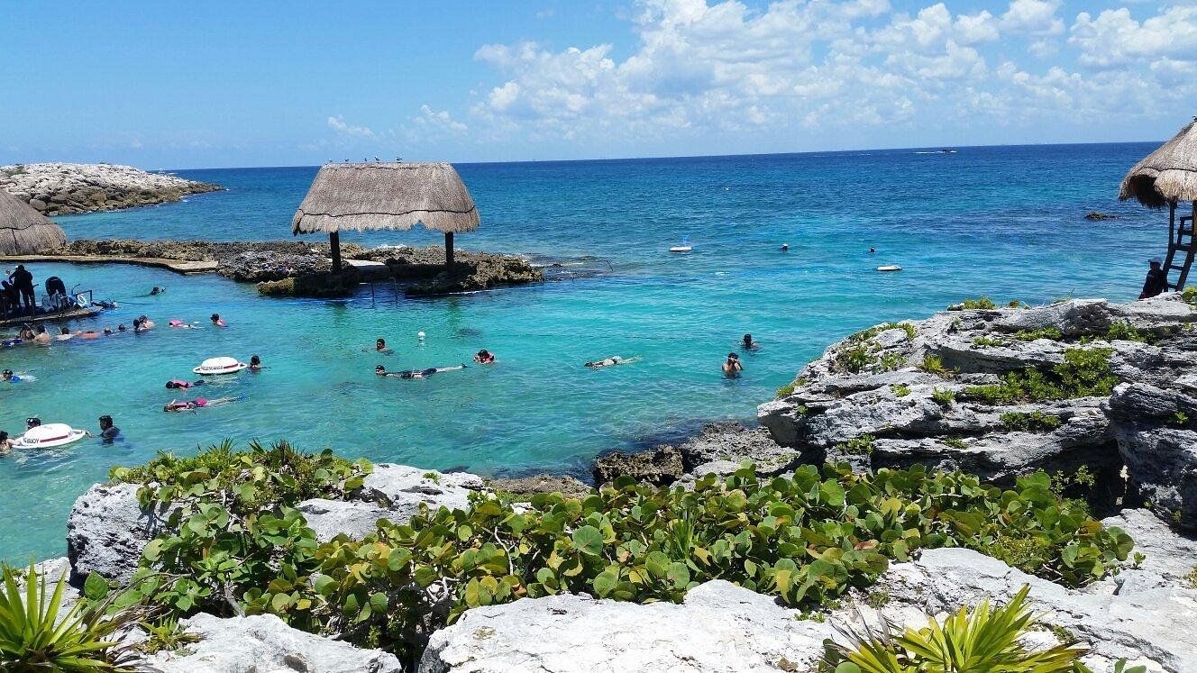 Escena de playa tropical con agua turquesa clara, gente nadando y un refugio con techo de paja.